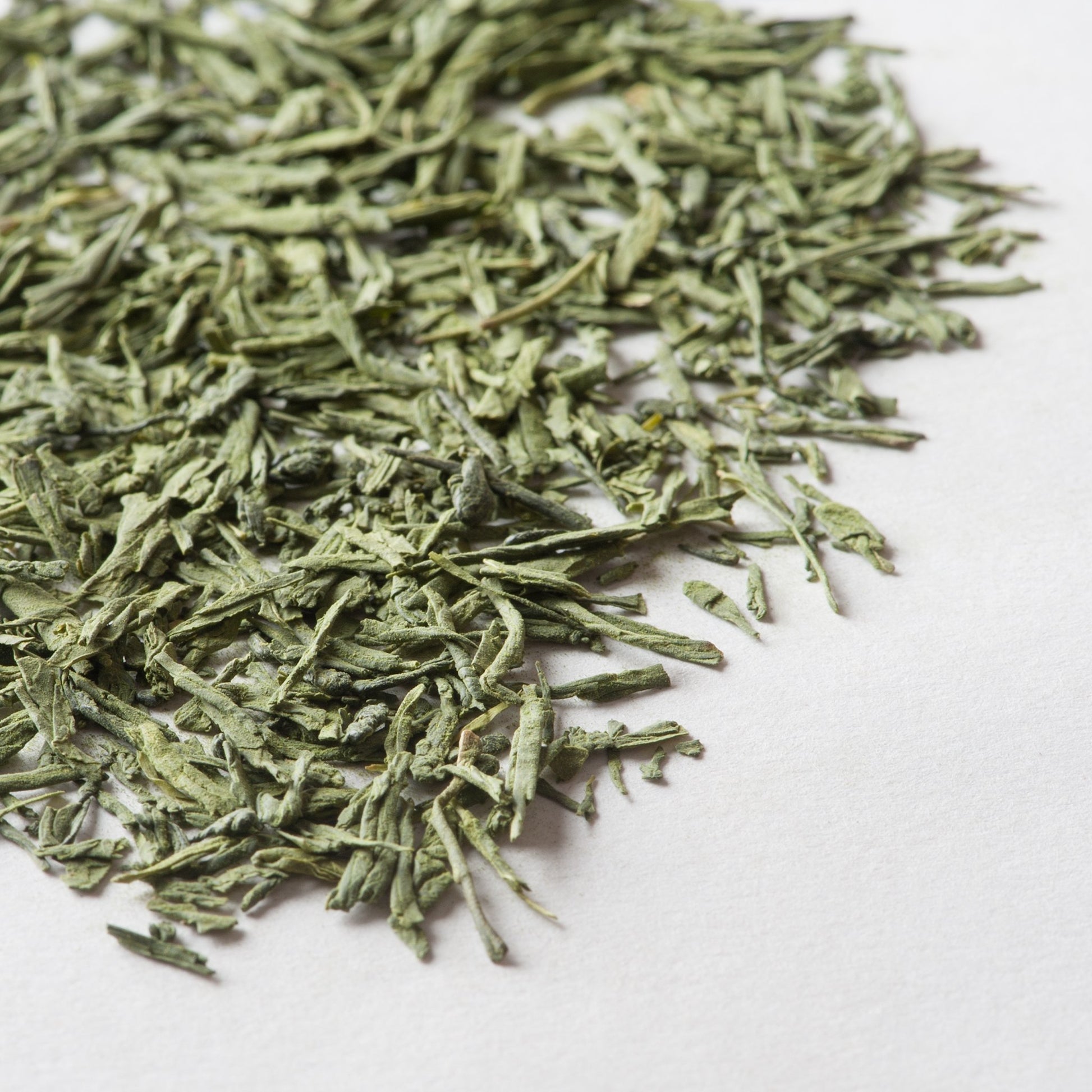 Loose green tea leaves with thin, needle-like strands in pale and dark green tones on white background.